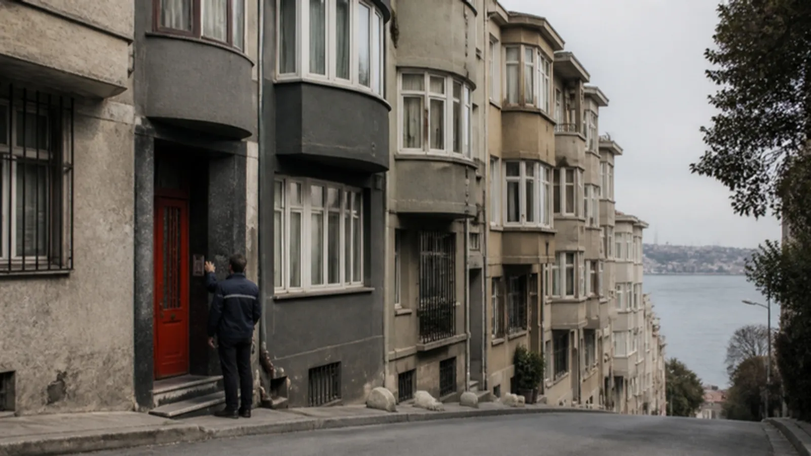Yokuşlu bir sokakta cumbalı eski apartmanlar ve girişini kontrol eden bina görevlisi