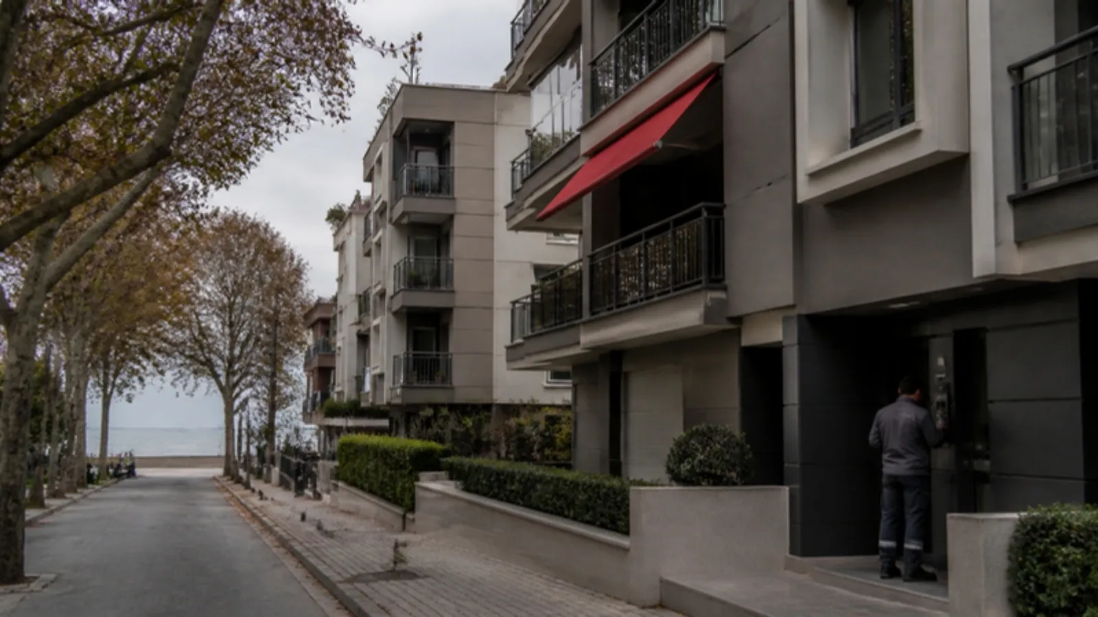 Deniz kıyısına yakın ağaçlı bir caddede orta katlı apartmanlar ve girişi kontrol eden teknisyen